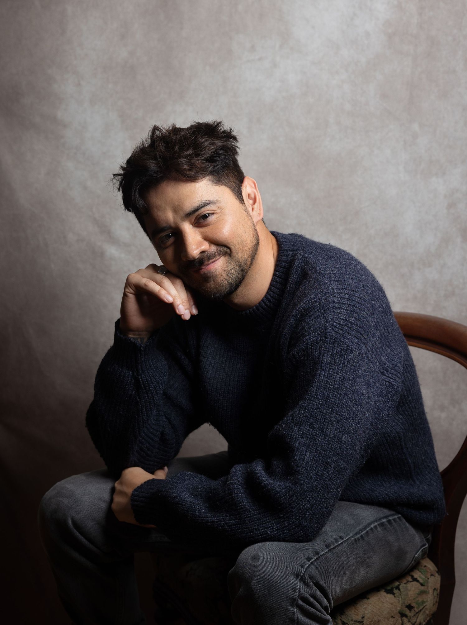 Smiling man in navy knit sweater sitting in wooden chair against neutral gray backdrop.