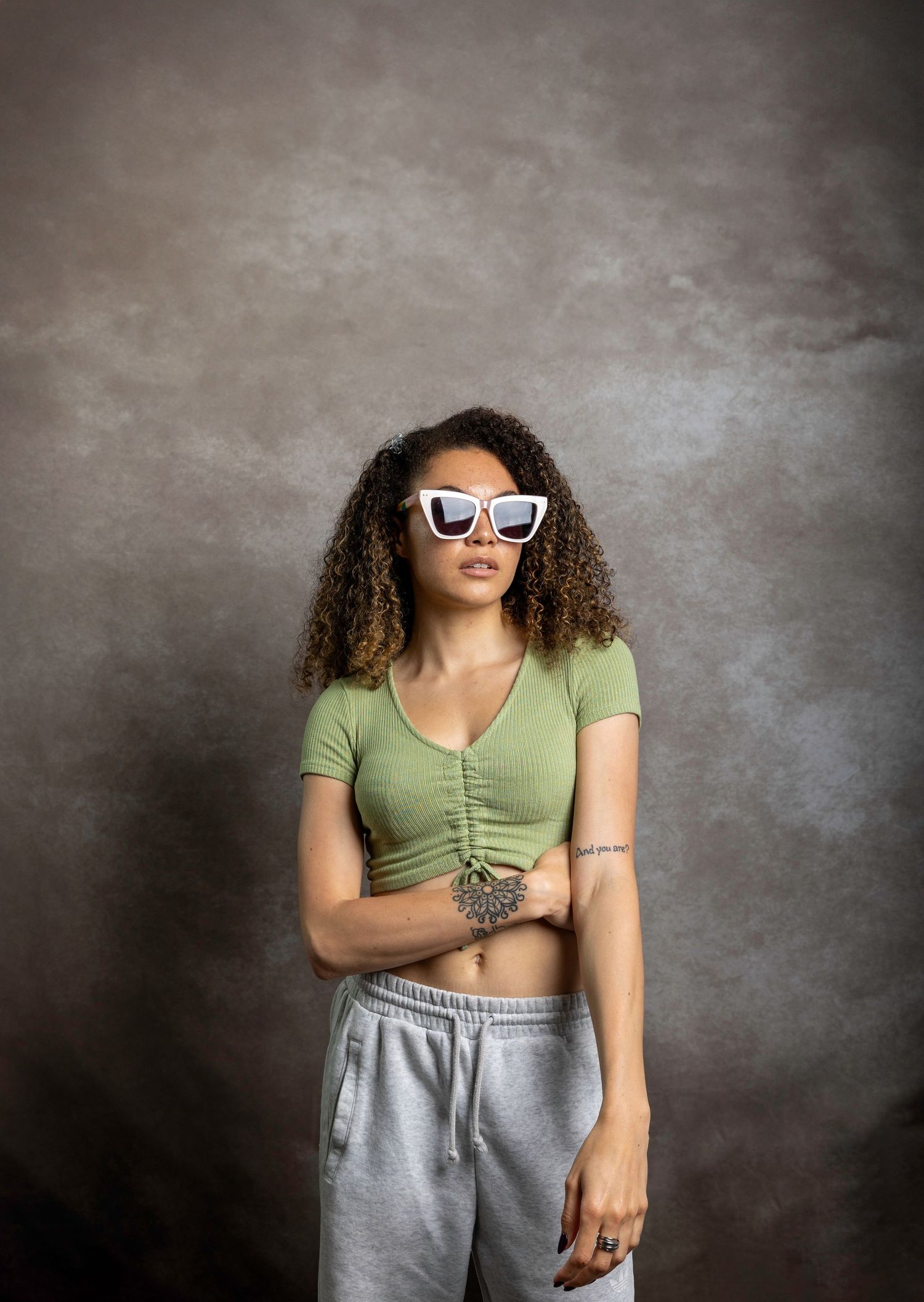 Woman in green crop top and grey sweatpants wearing white sunglasses, arms crossed in studio.