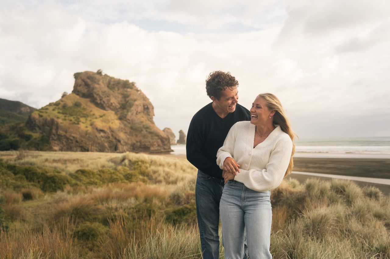 Couple smiling at each other in coastal grass with dramatic rock formation and beach behind them.