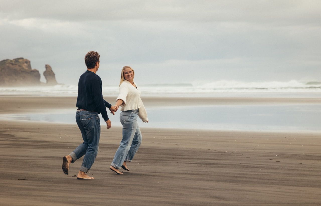 A couple holds hands and walks joyfully on a dark sandy beach with ocean waves and misty sea stacks in the background.