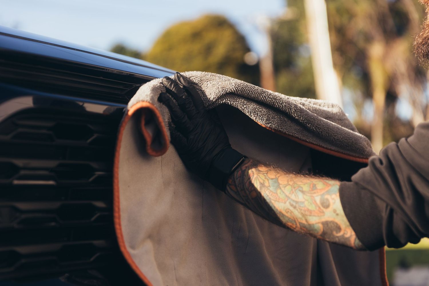 Close-up of a tattooed hand wearing black gloves wiping a dark vehicle with a microfiber cloth outdoors.
