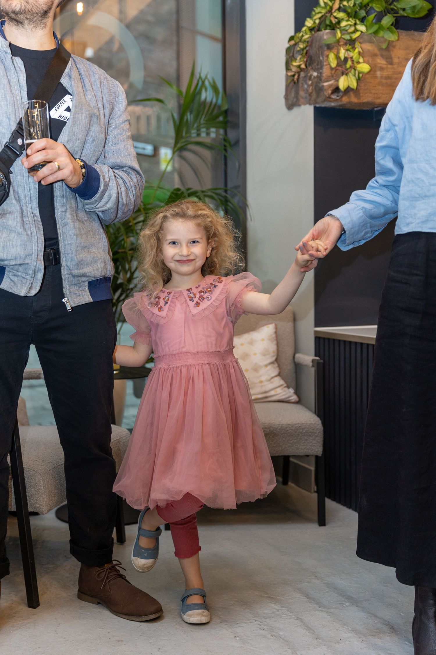 Adorable curly-haired girl in a pink dress holding hands with adults at an indoor event.