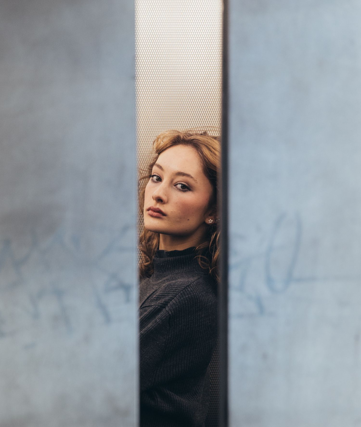Young woman with auburn hair peering through narrow gap between two metal panels.