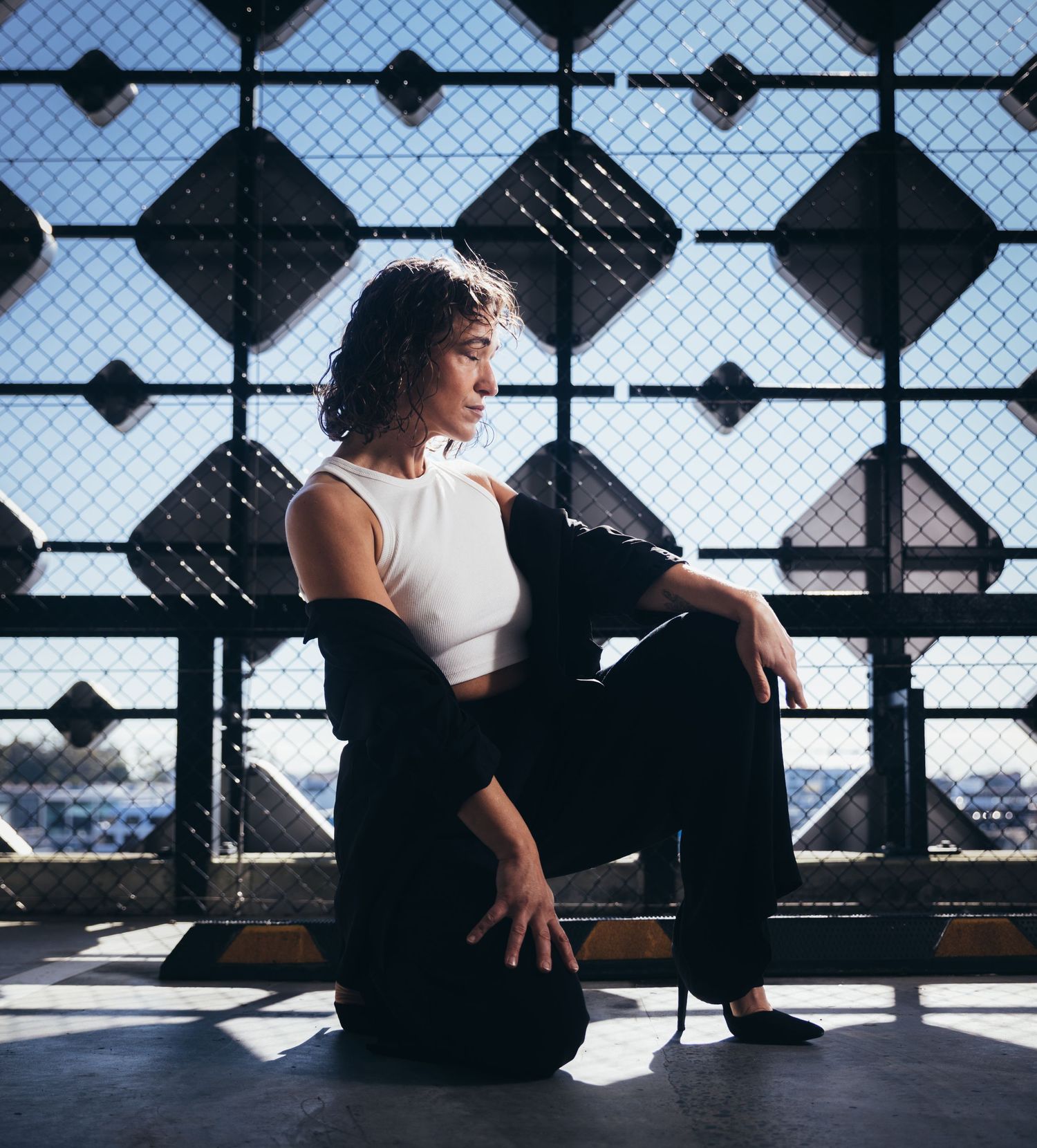 Woman in white crop top and black pants sitting pensively against geometric patterned metal fence.