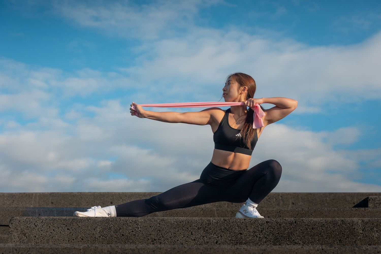 Fit woman in black activewear performing a lunge with resistance band against a blue sky.