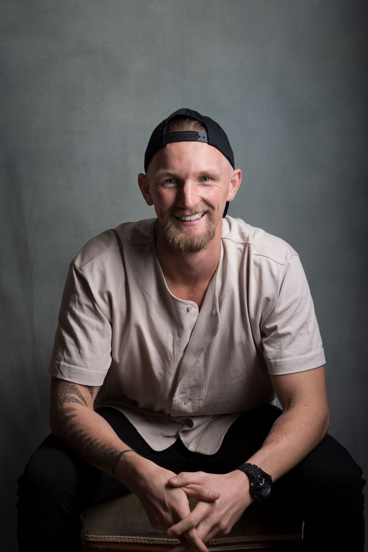 Smiling man in beige shirt sitting with hands clasped wearing backwards baseball cap.