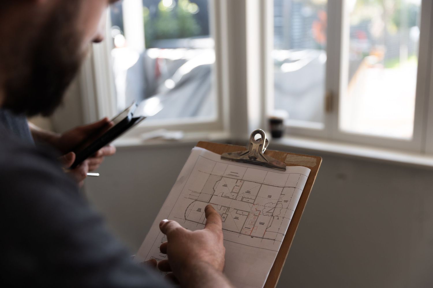Bearded builder reviewing detailed floor plan blueprints on a clipboard near a window.