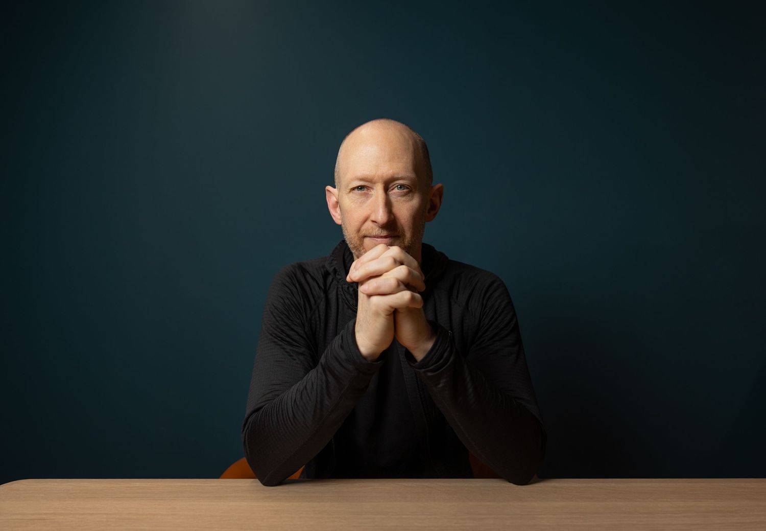 Bald man in a black turtleneck with hands clasped seated at a wooden table against a teal wall.