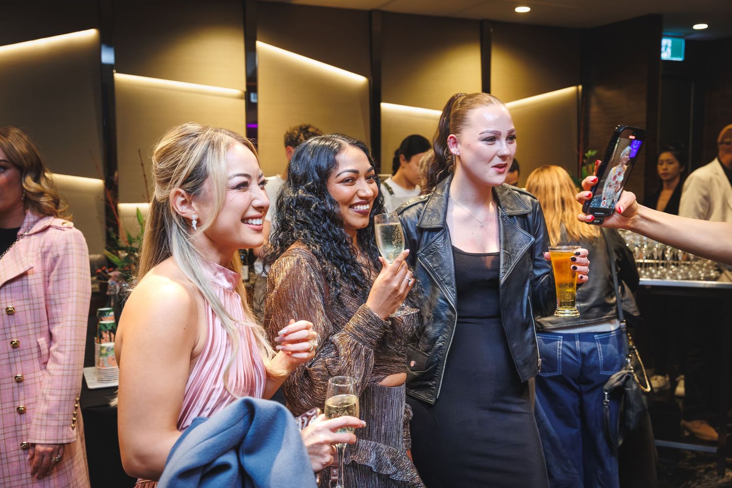 Three stylish women laughing and holding champagne glasses at a lively indoor social event.