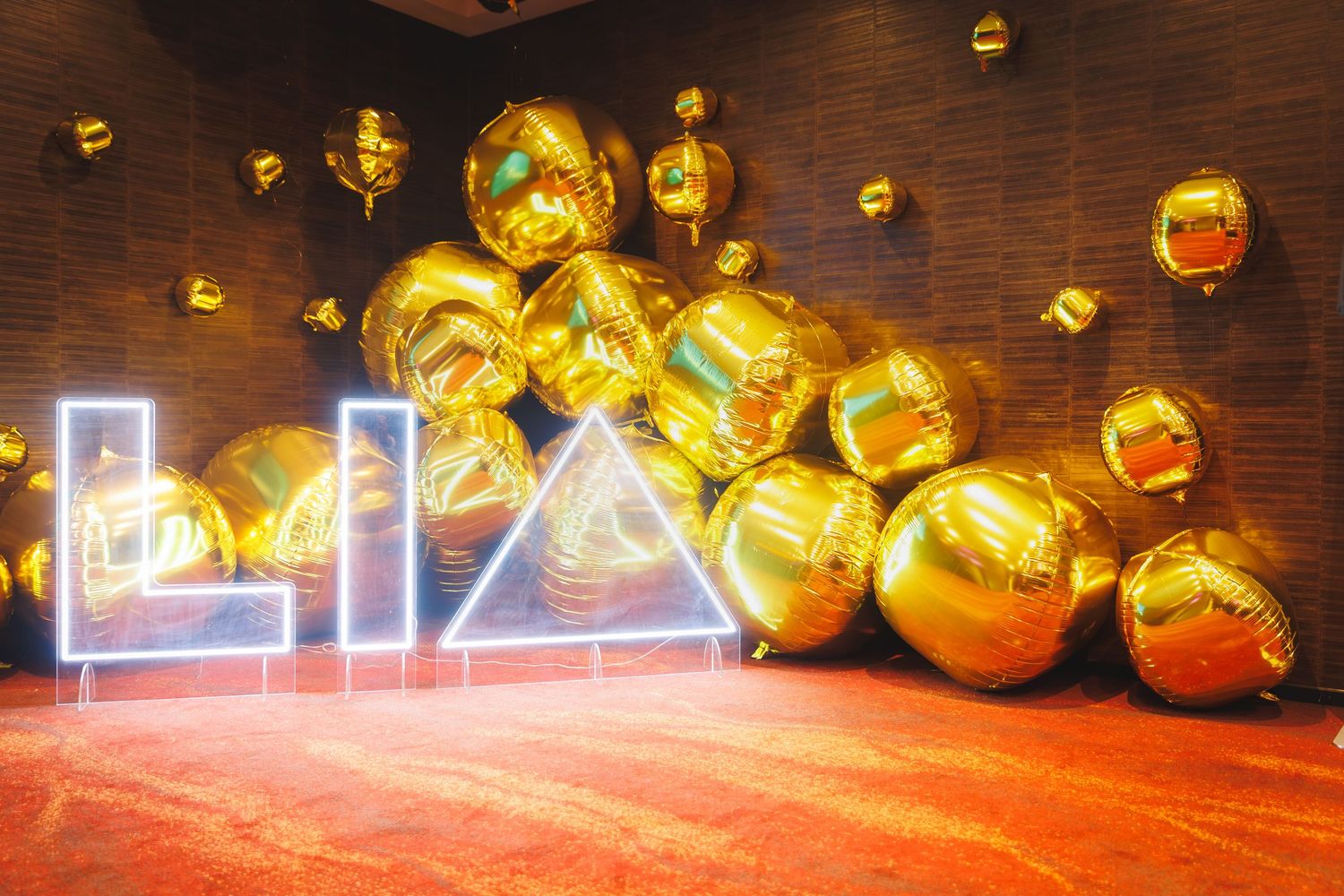 Gold metallic balloons surround a neon LIA sign installation against a brick wall backdrop.
