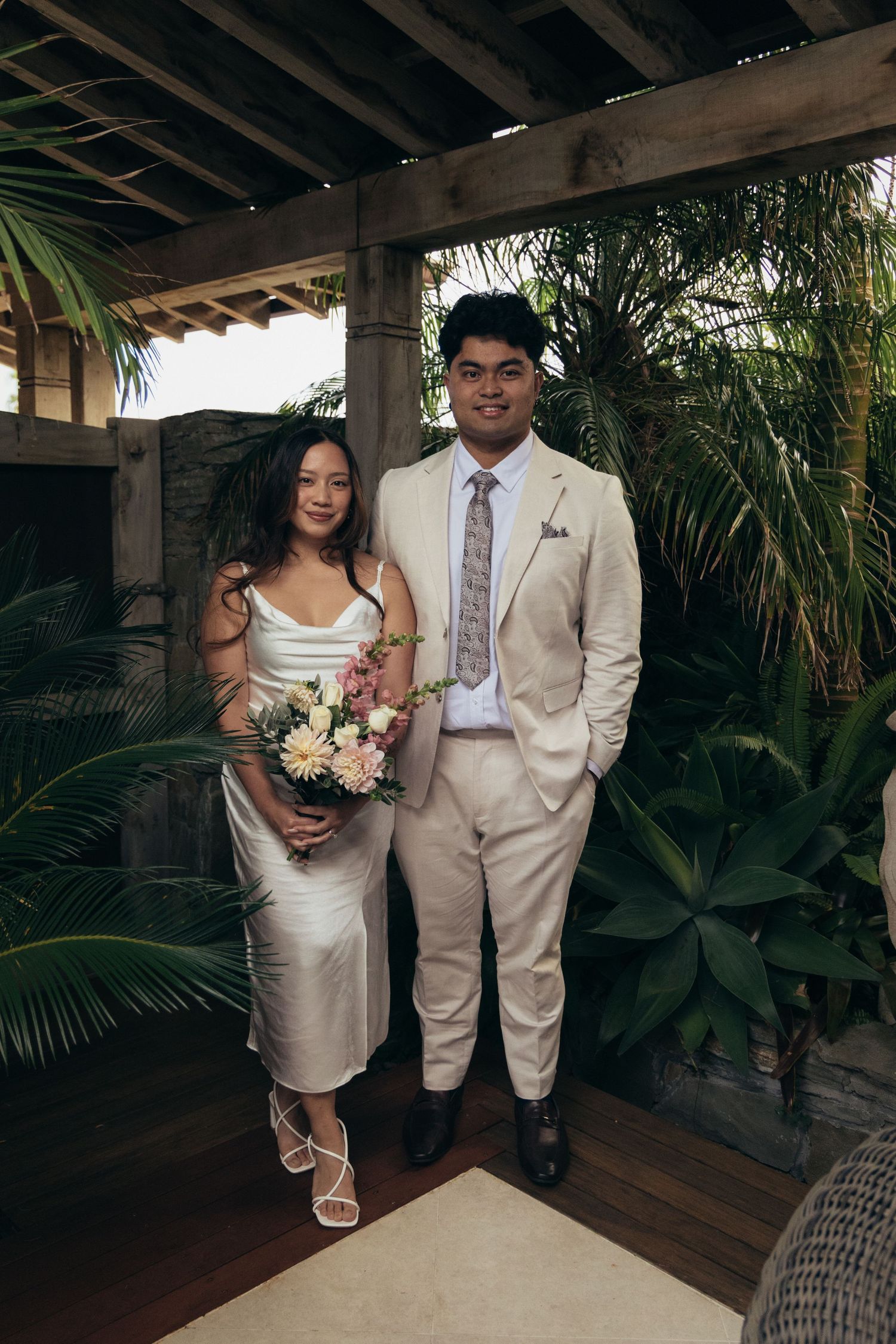 Couple posing in tropical garden setting, woman in silver dress holding bouquet, man in beige suit with floral tie.