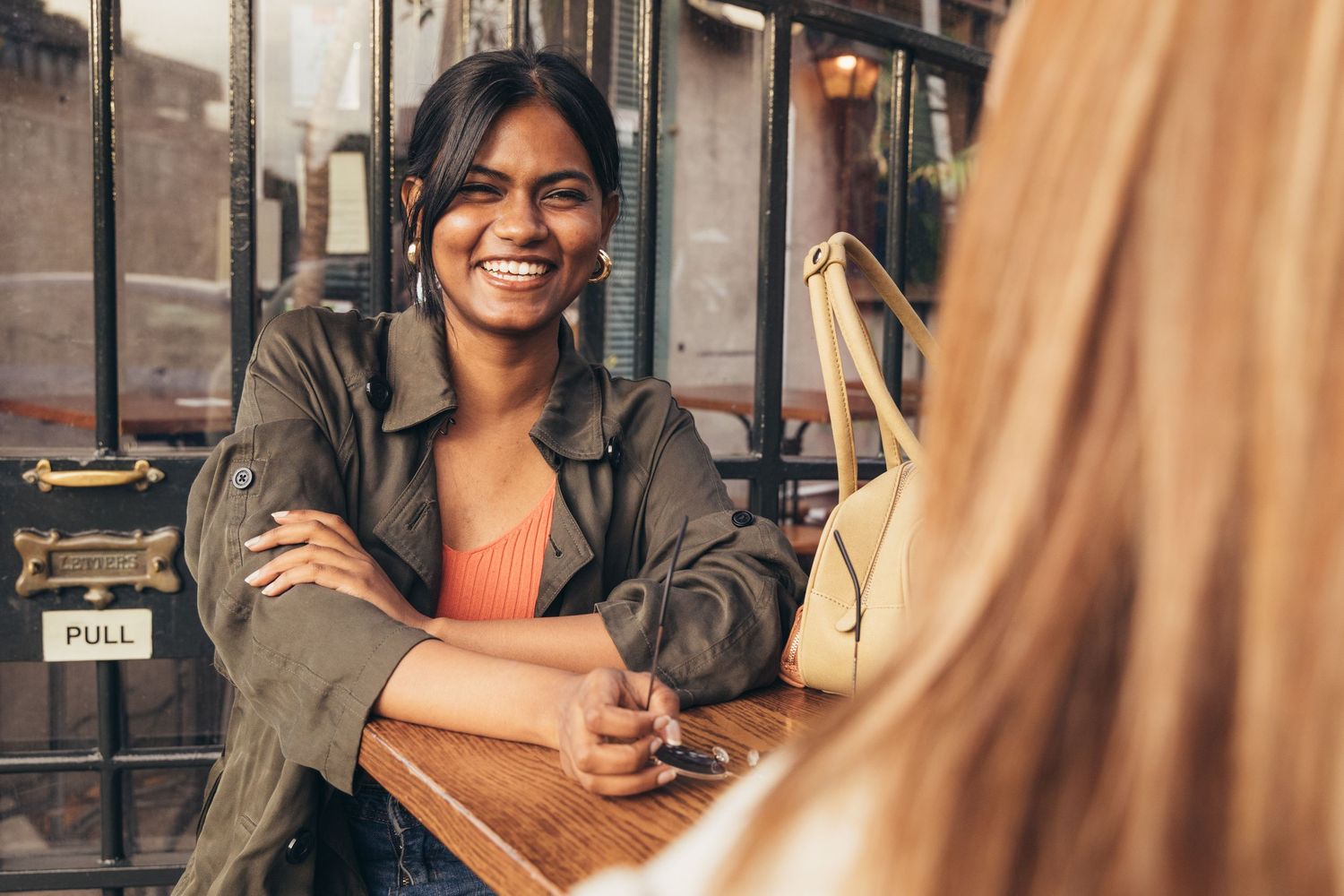 Two women smiling and chatting at an outdoor cafe table, one with crossed arms wearing a green jacket.