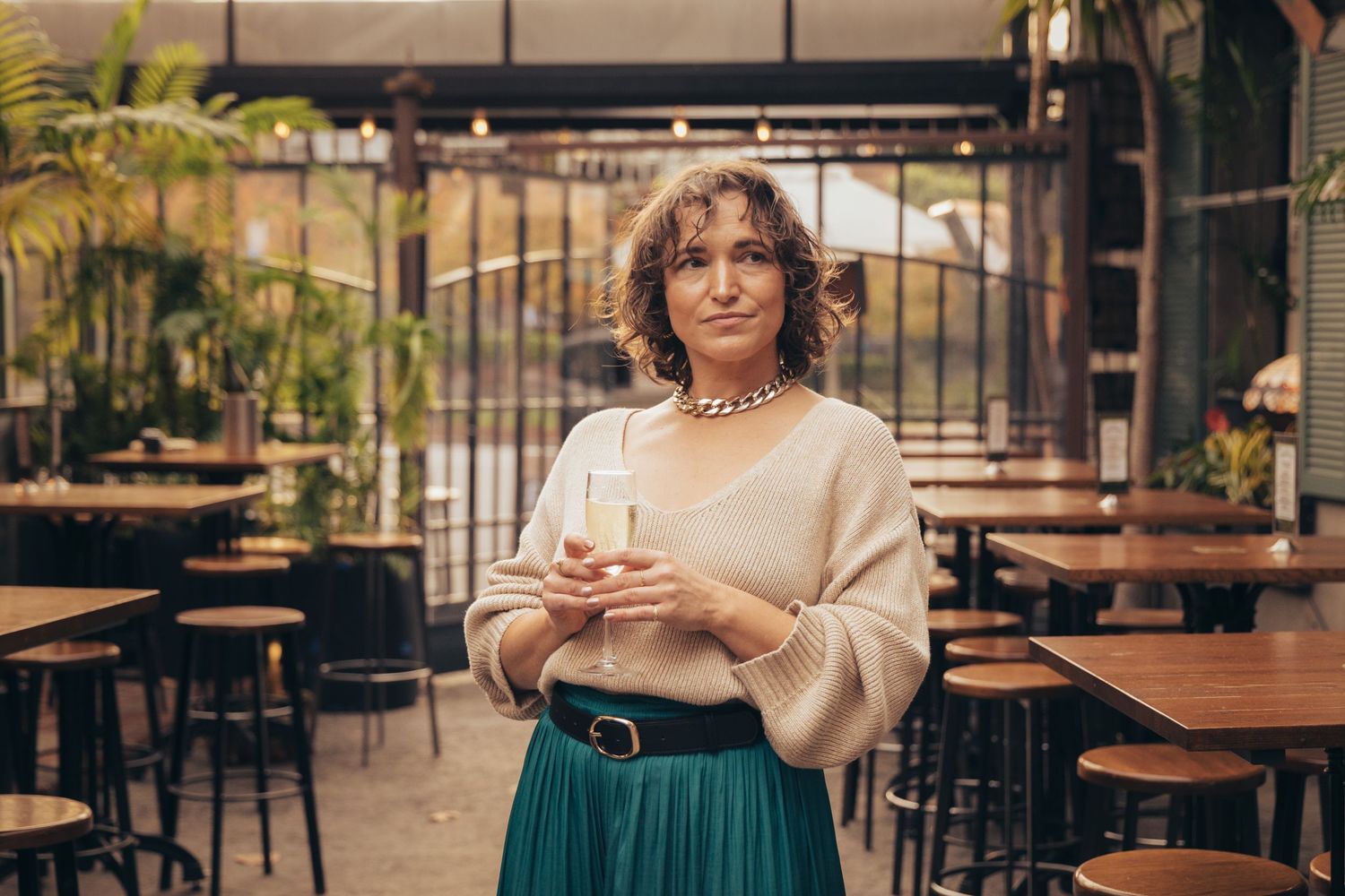 Woman in teal skirt and beige top holding champagne glass in stylish outdoor restaurant with tropical plants.