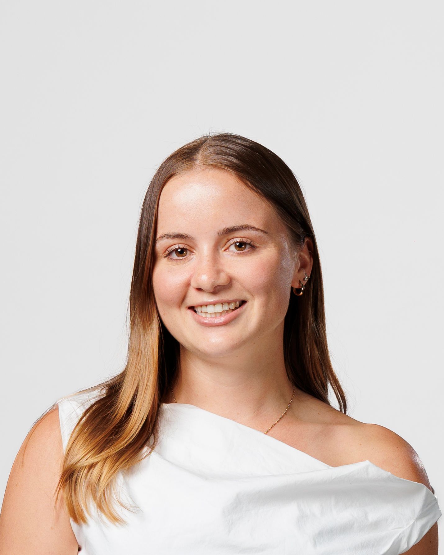 Young woman with long brown hair smiling, wearing a white one-shoulder top against a light grey background.
