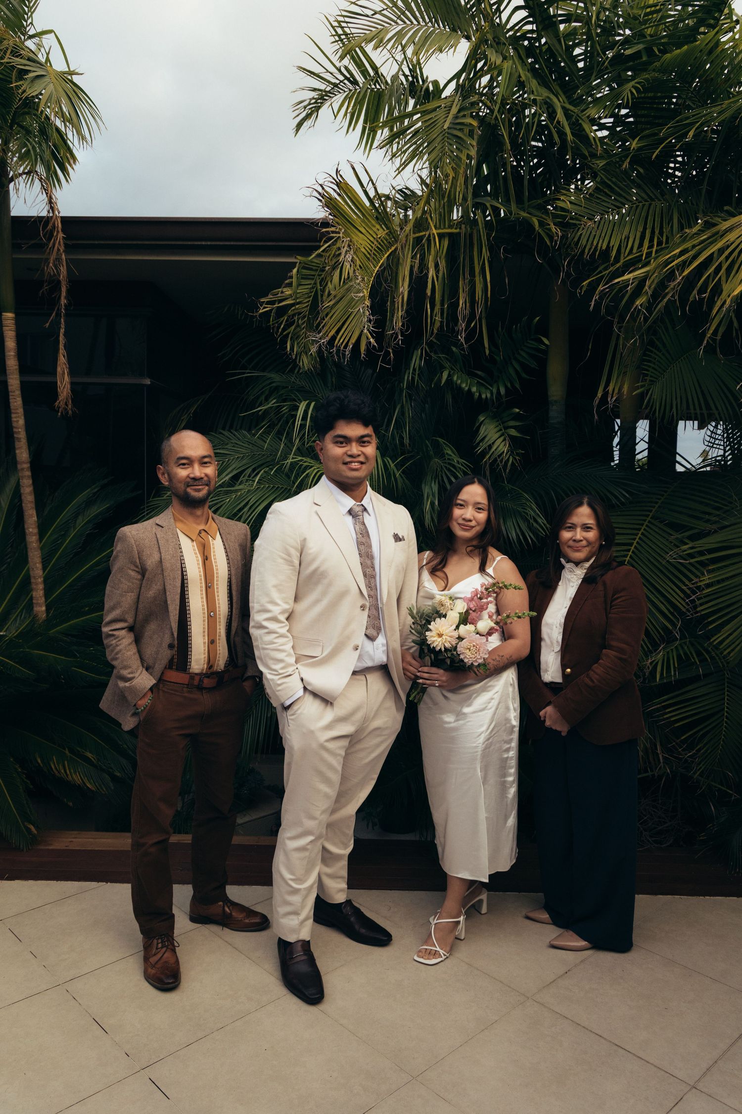 Wedding party of four posing outdoors with tropical palms, bride in white dress holding bouquet, groom in cream suit.