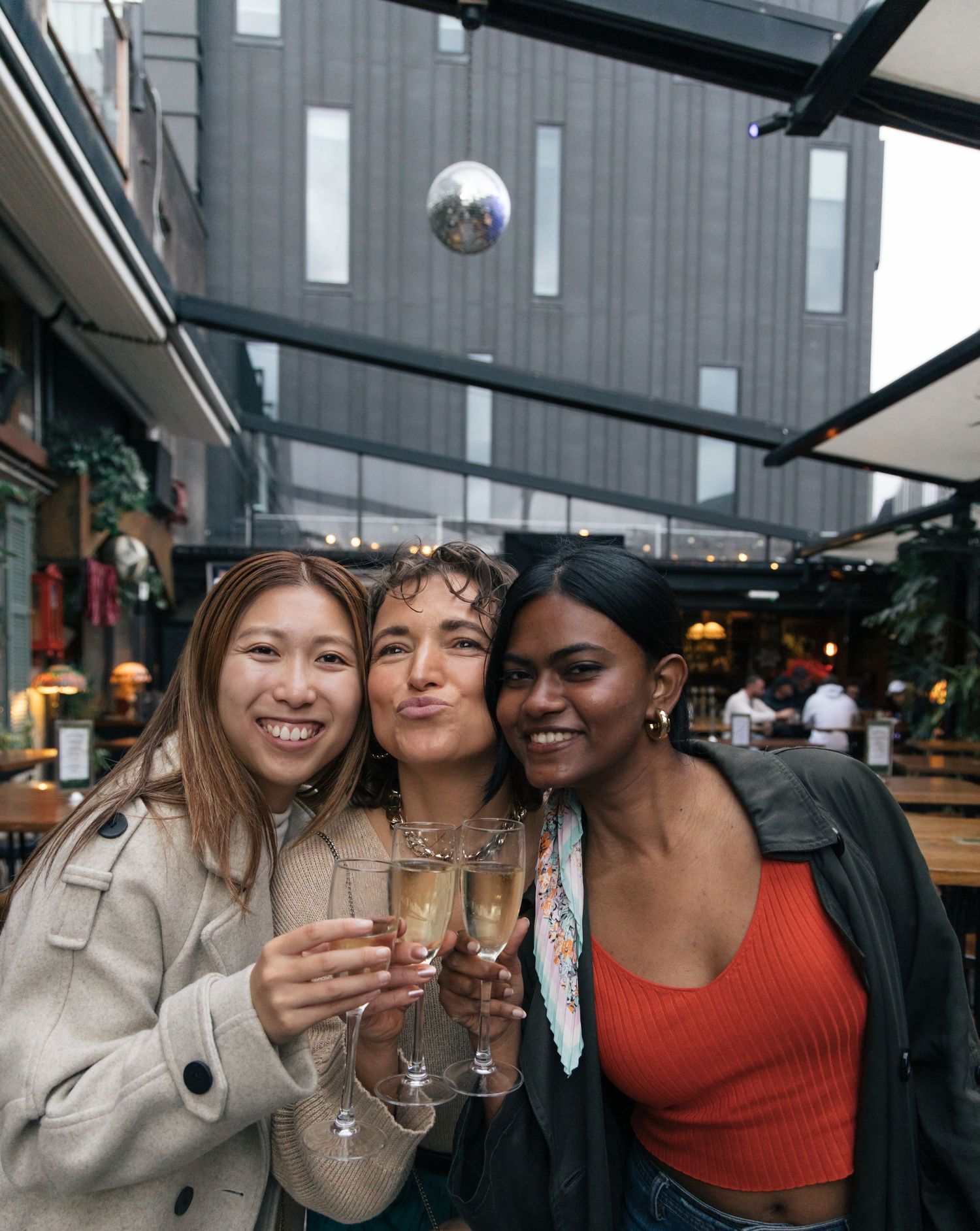 Three smiling diverse female friends toasting with champagne flutes at an outdoor bar.