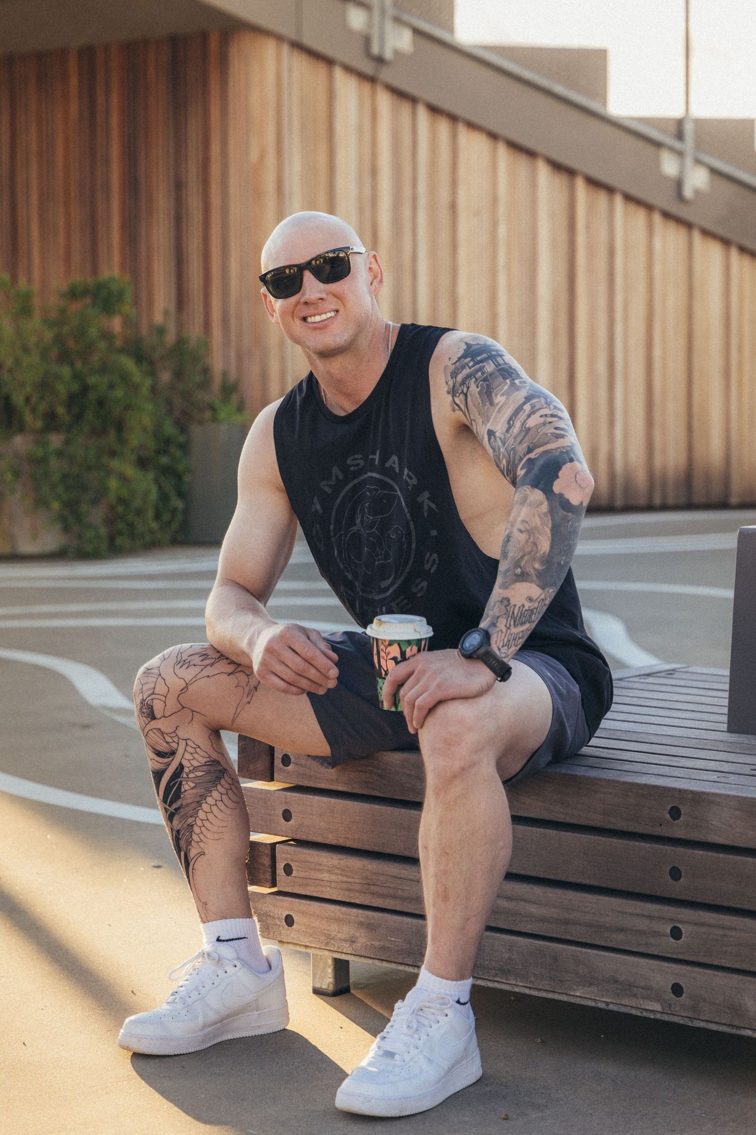 Smiling tattooed bald man sitting on bench holding coffee cup wearing sunglasses and Nike sneakers.