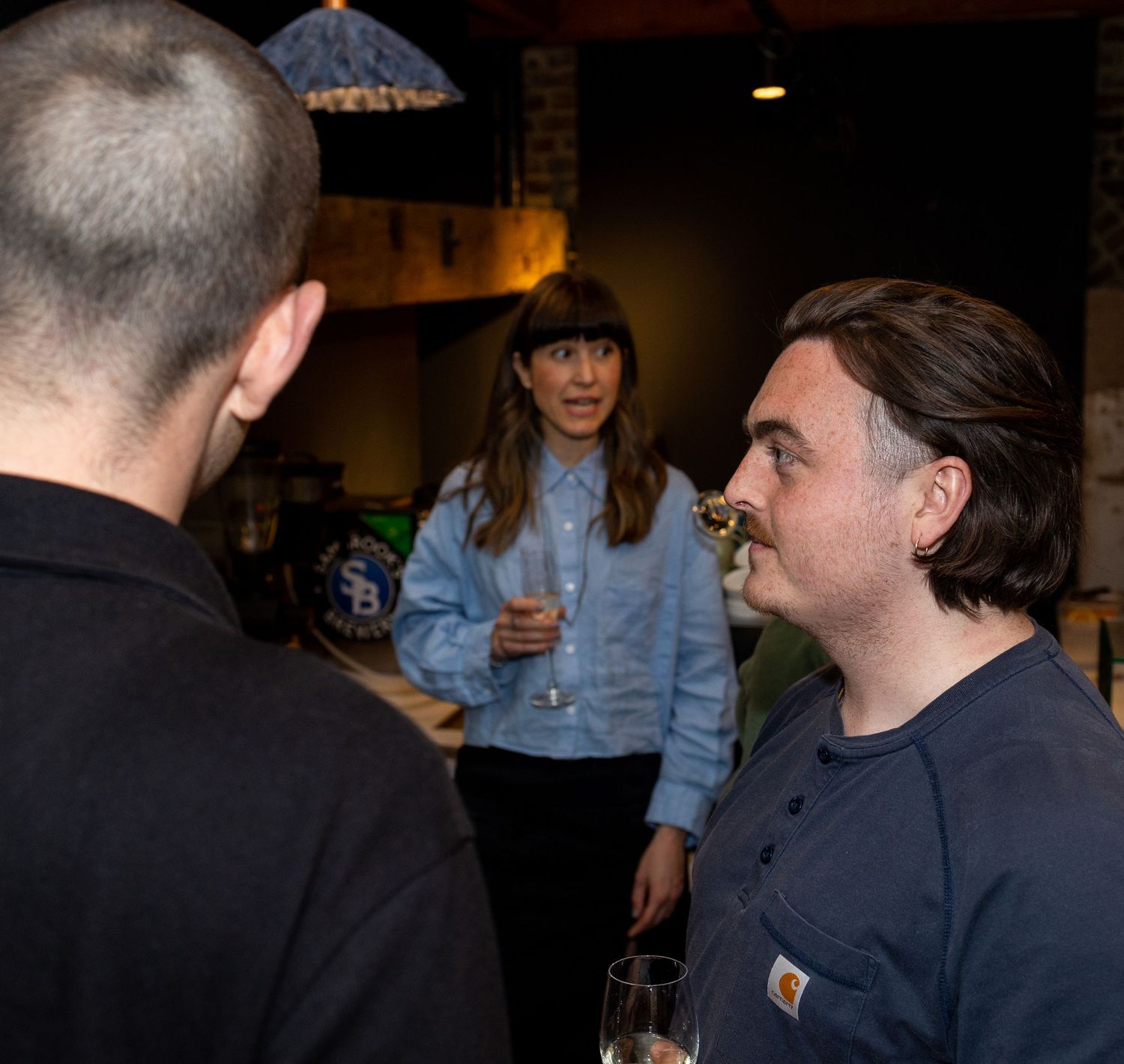 Three people engaged in conversation at a casual indoor gathering, holding wine glasses.