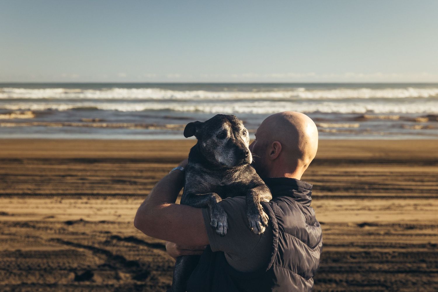 Bald man holding black dog up to his face on sandy beach with ocean in background.