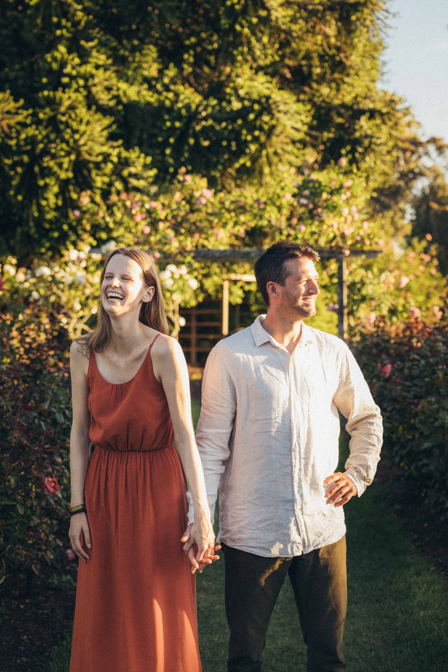 A happy couple holds hands and laughs while standing in a beautiful sunlit garden with roses.