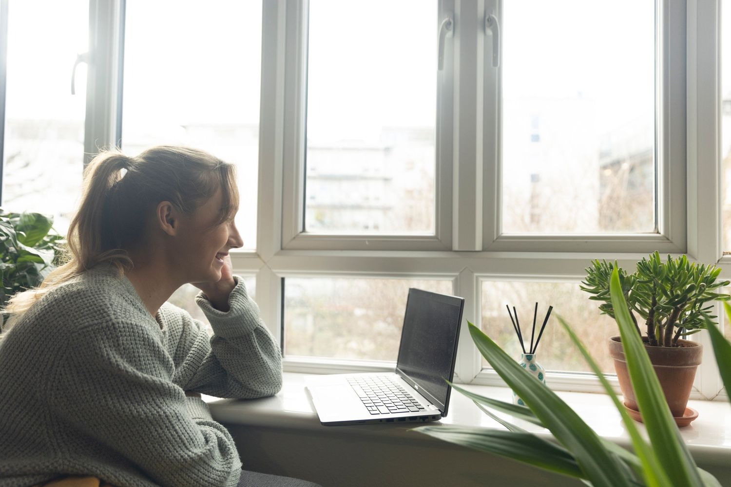 Woman in gray sweater sitting by window looking outside with open laptop on desk beside plants.