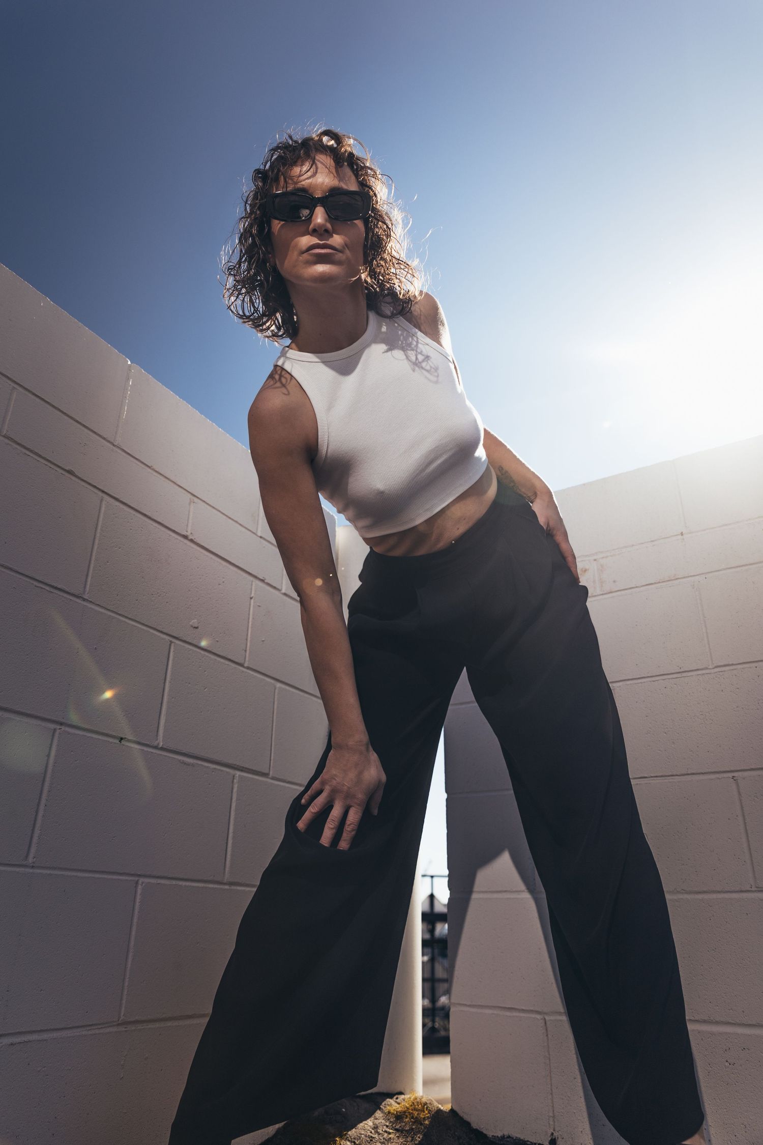 Woman in white crop top and black wide-leg pants leans against white brick wall in bright sunlight.
