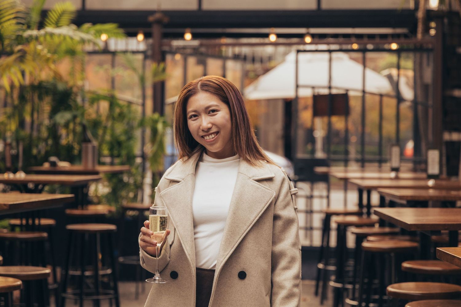 Smiling Asian woman holding a champagne glass in a stylish outdoor restaurant.