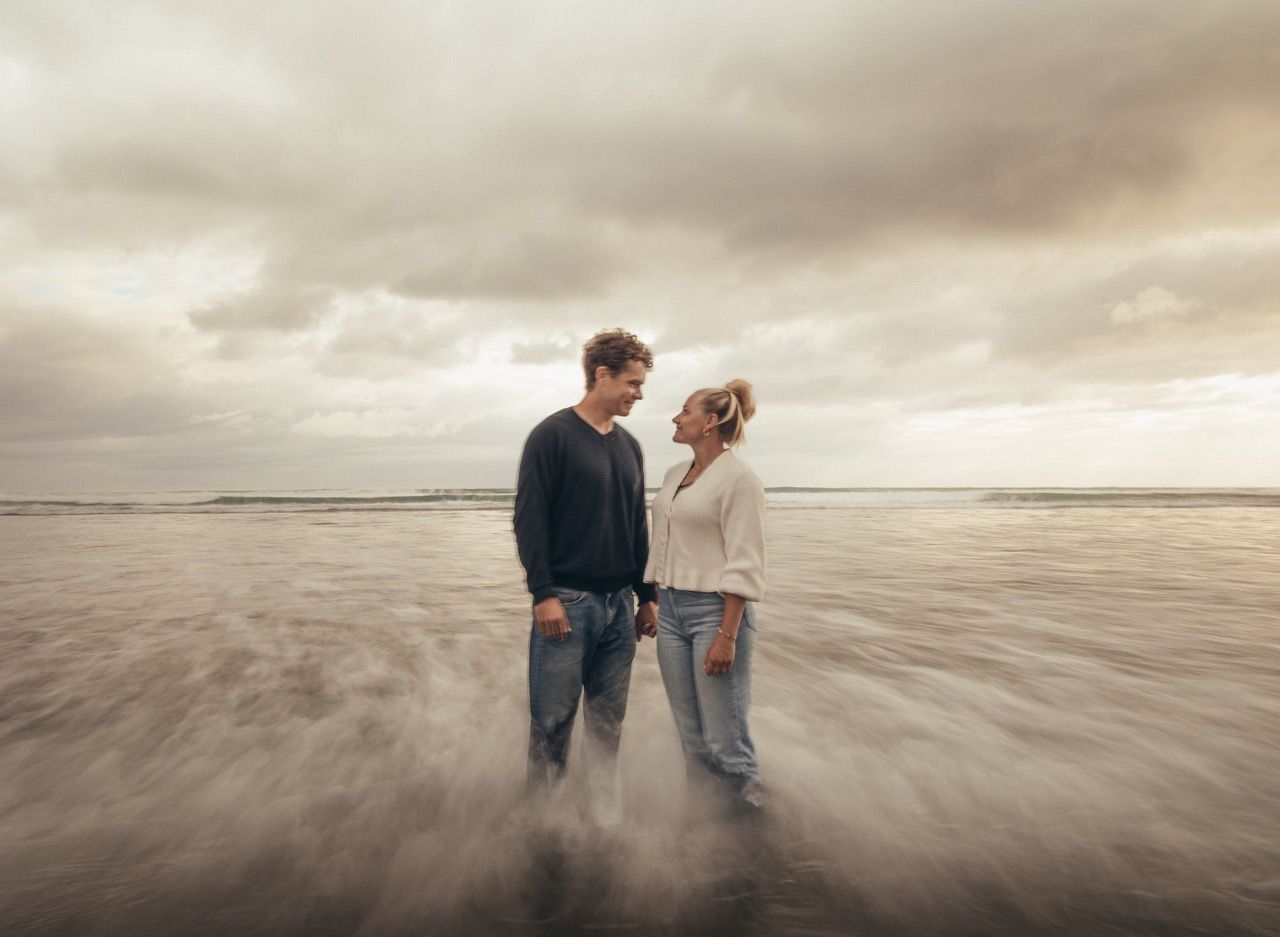 Couple standing face to face on a misty beach with dramatic cloudy skies and ocean waves surrounding them.