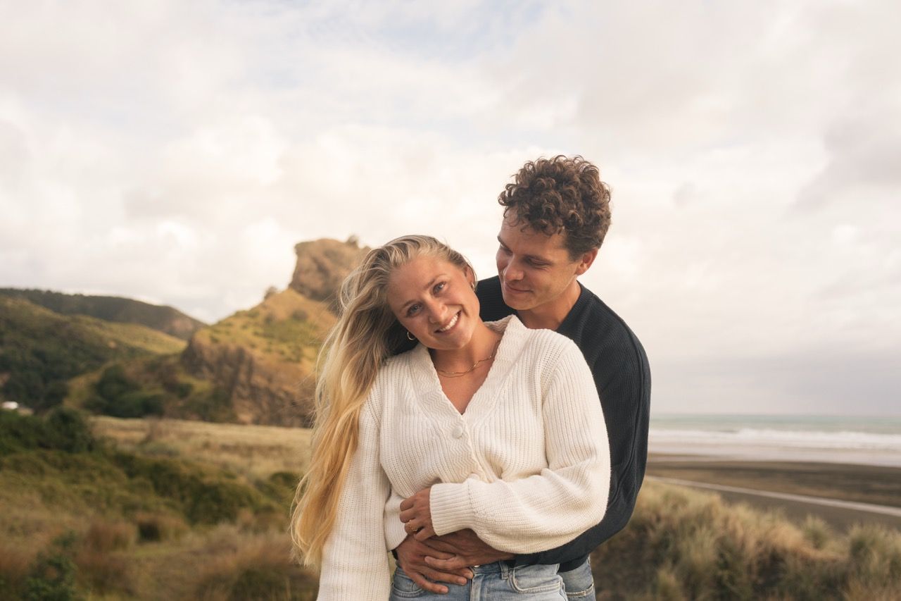 A couple embraces on a coastal hillside, the woman smiling warmly, with a beach and rocky cliffs in the background.