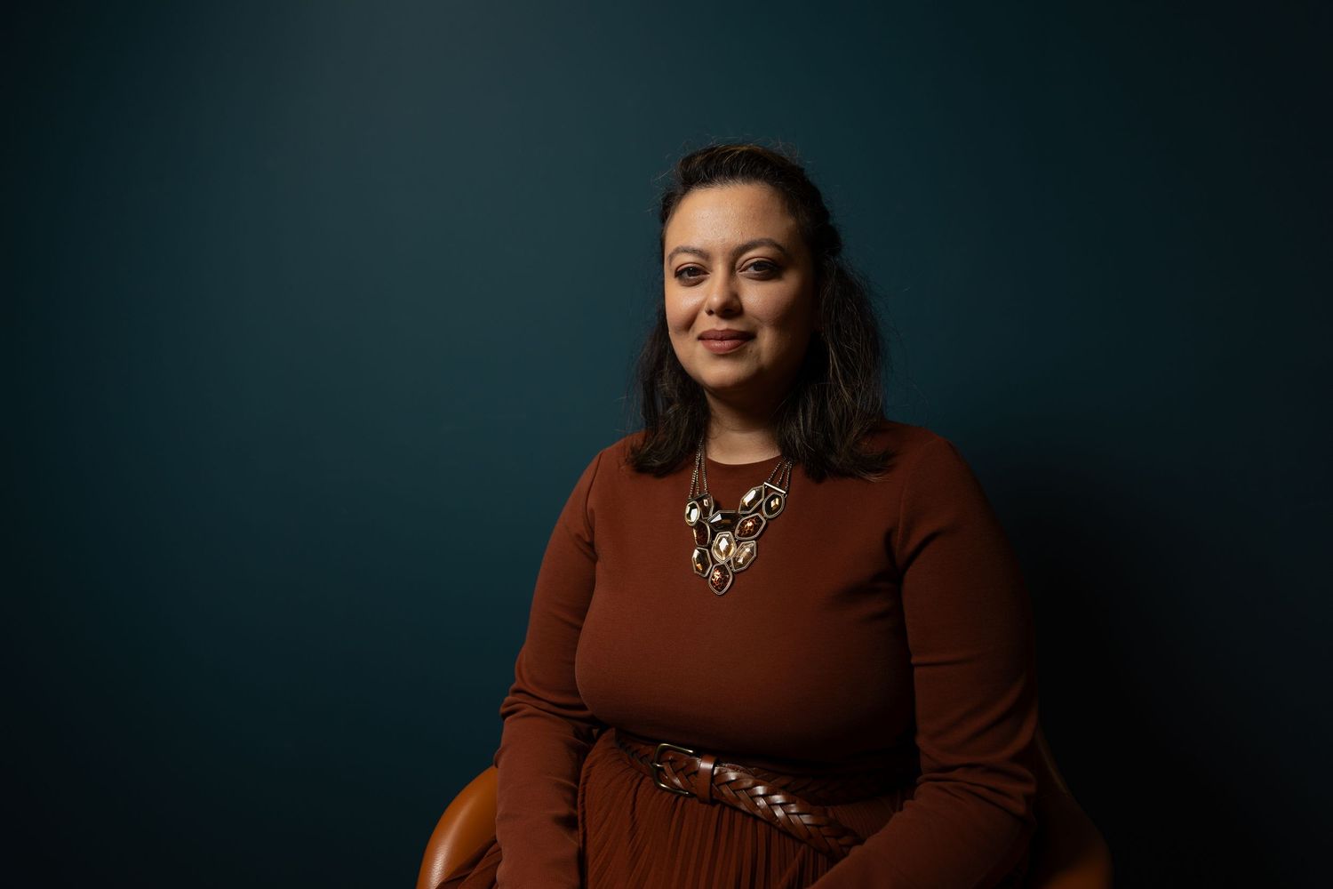 Smiling woman in a rust-colored top with a gold necklace seated against a dark teal background.