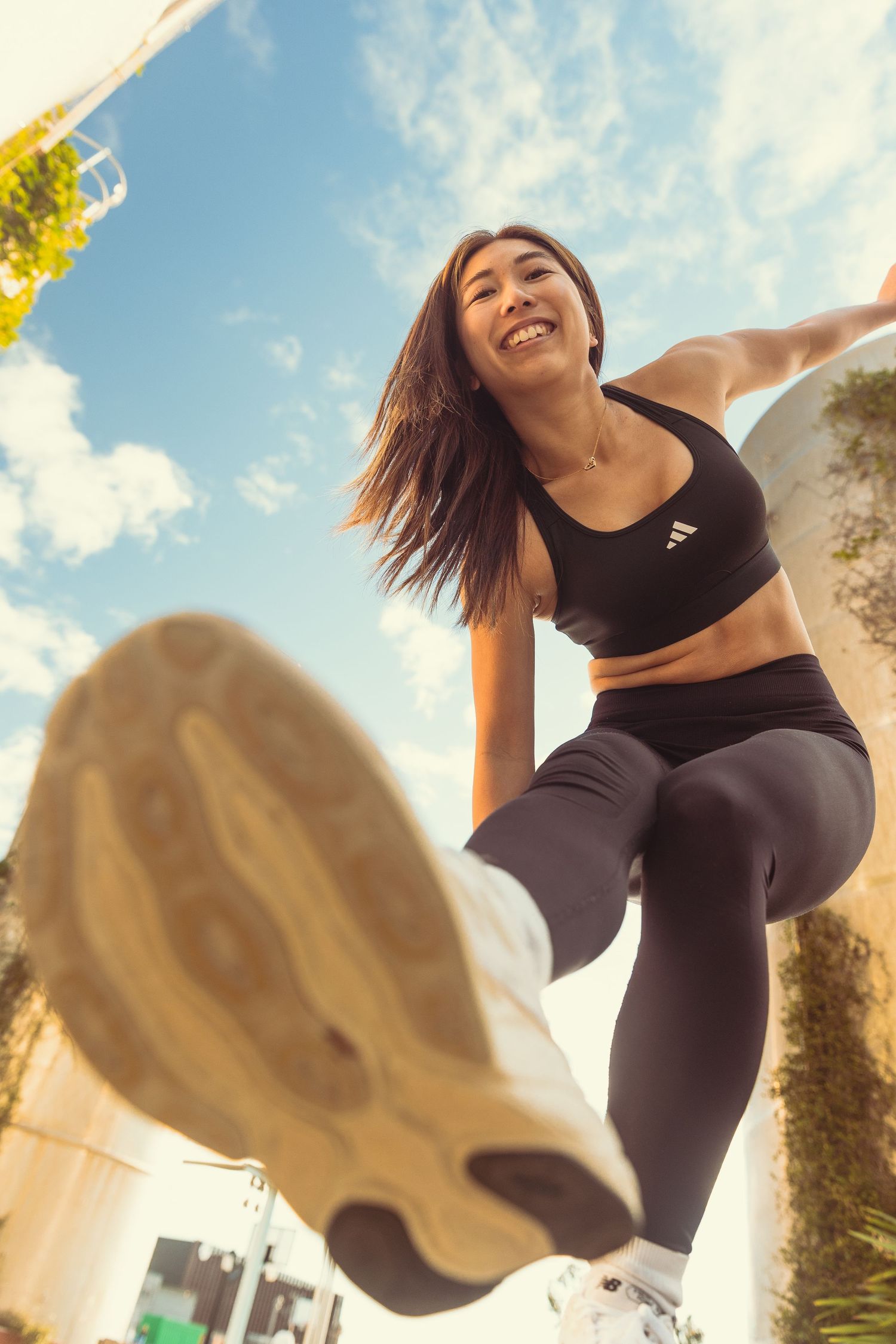 Smiling woman in black Adidas sports bra and leggings doing a high knee exercise outdoors under a blue sky.