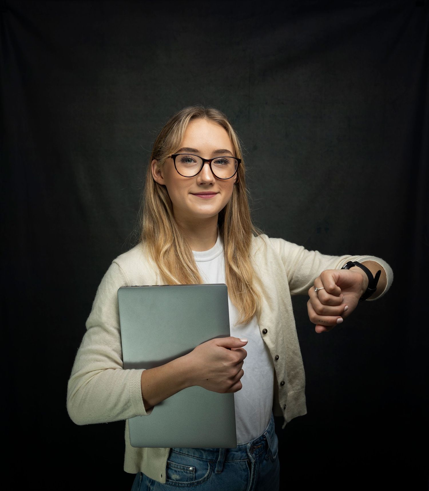 Young blonde woman in glasses holding laptop and checking smartwatch against dark studio background.