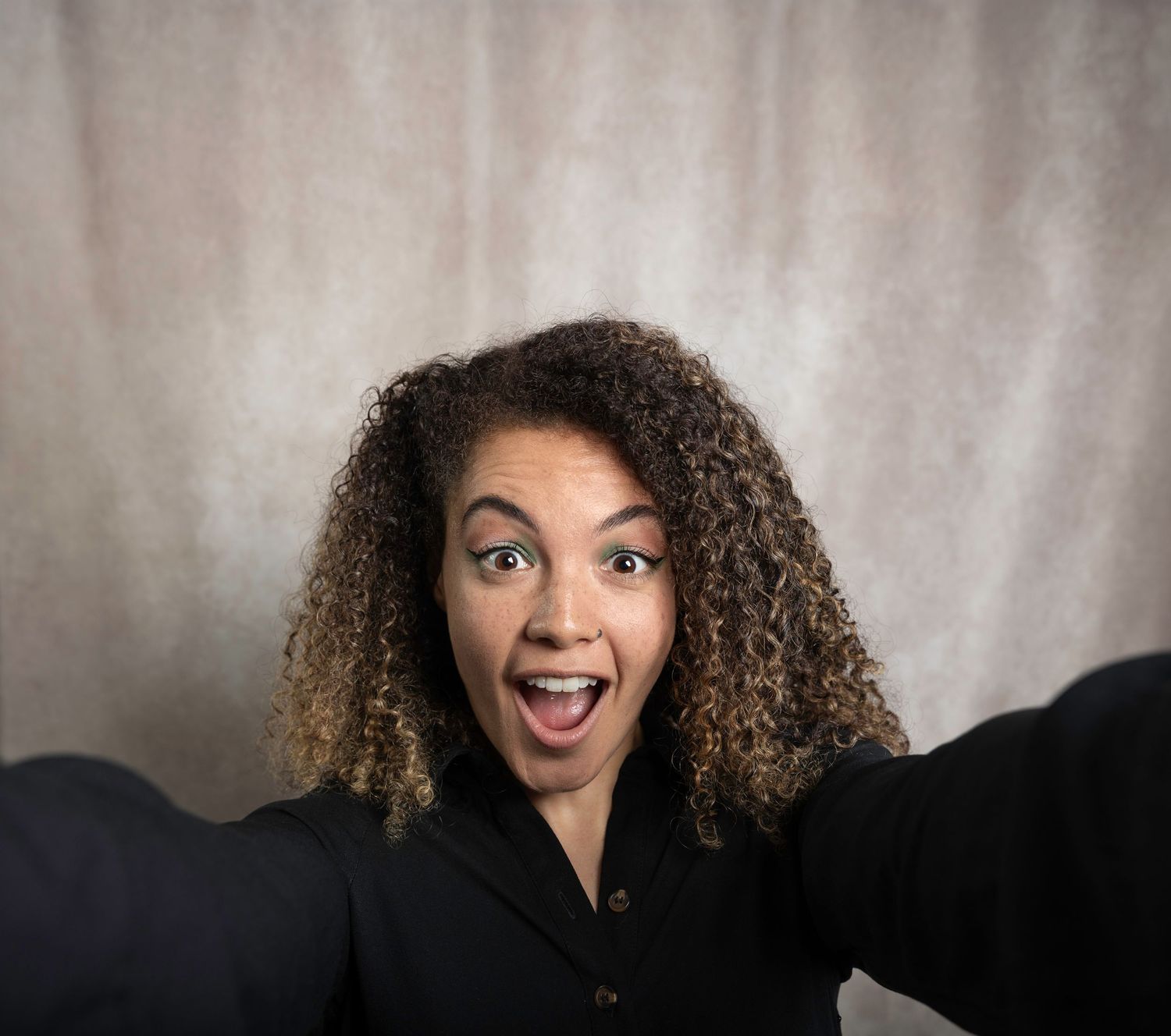 Excited woman with curly hair taking selfie, mouth open wide, wearing black blazer.