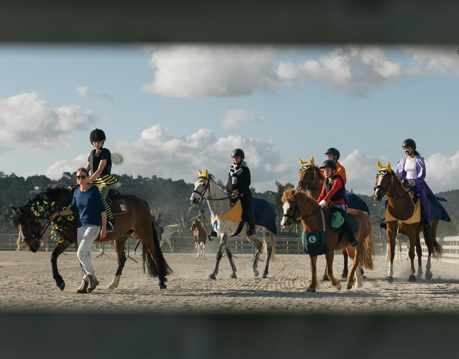 Group of five equestrians on horses parading through sandy arena under partly cloudy sky.