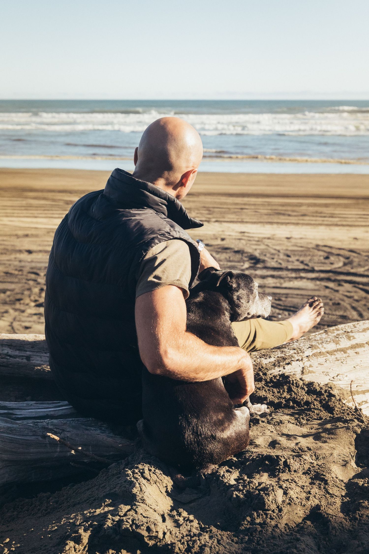 Bald man sits on driftwood embracing his black dog while gazing at ocean waves.