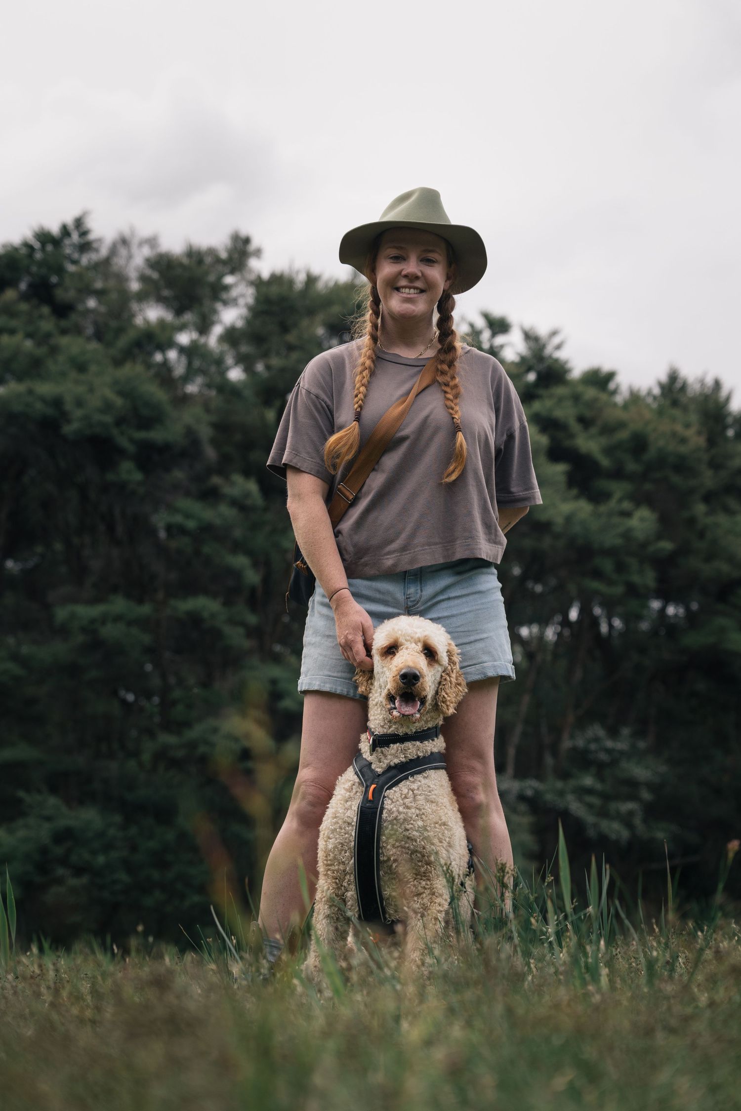 Smiling woman in wide-brim hat standing with golden labradoodle dog in grassy field near forest.