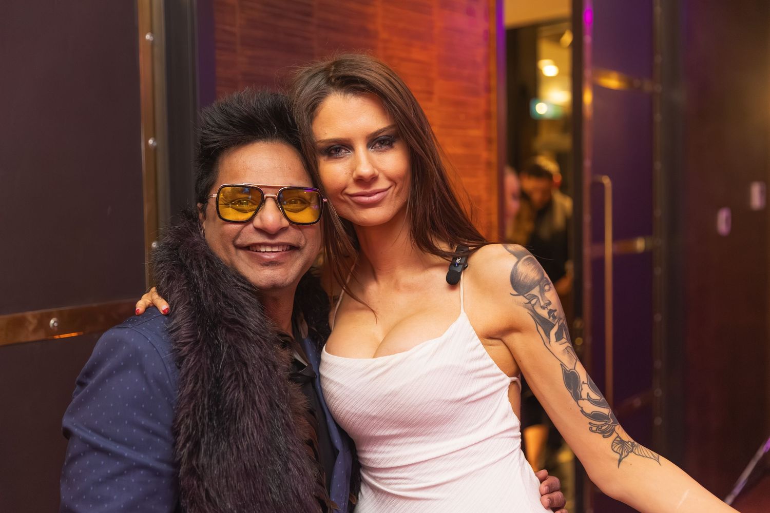 Two people smiling together at an indoor event, one wearing bold yellow sunglasses and fur jacket.