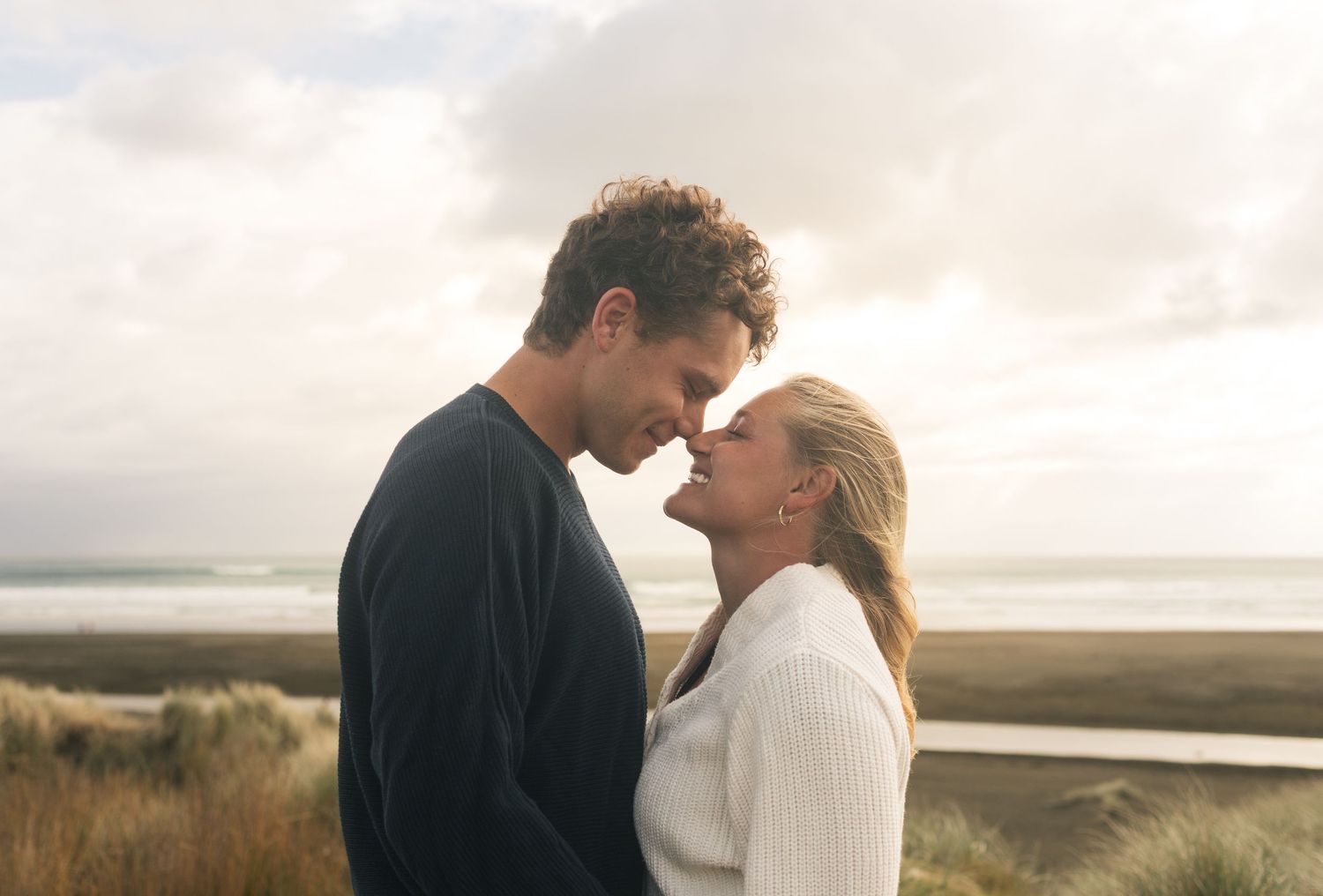 Romantic couple touching foreheads on a beach, sharing an intimate moment with ocean and cloudy sky behind them.