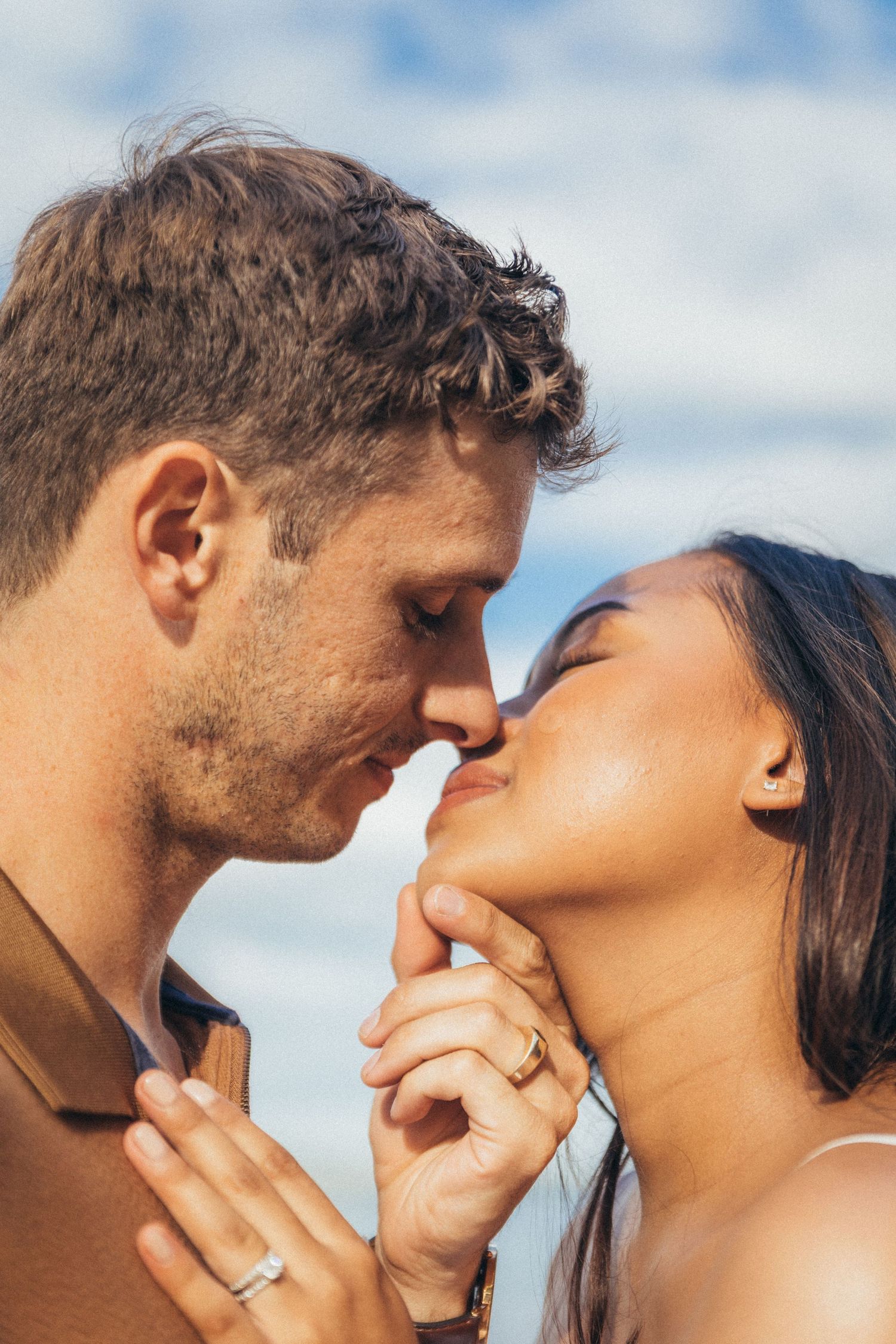 Romantic couple sharing an intimate moment, almost kissing outdoors, woman wearing engagement ring, blue sky background.