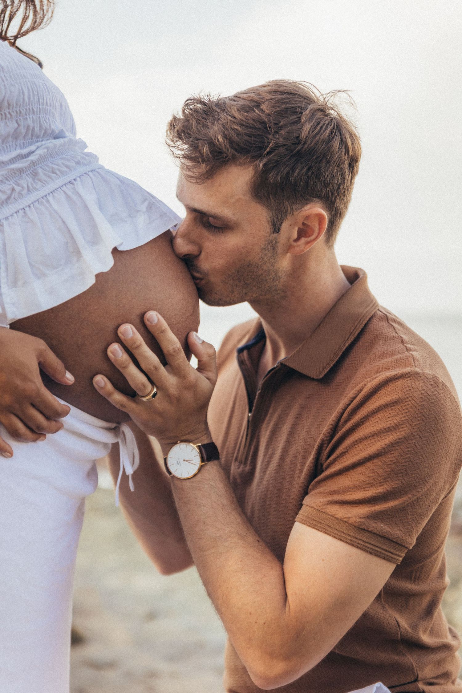 Man kissing pregnant woman's belly on beach, tender maternity moment with couple in casual summer outfits.
