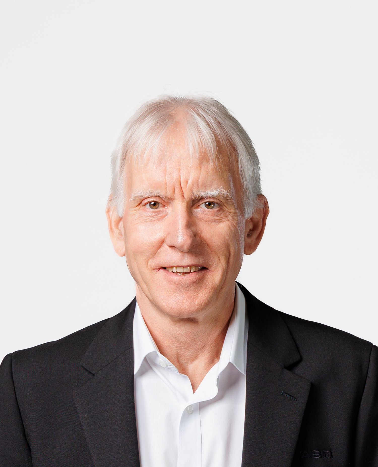 Older man with white hair wearing a black blazer and white dress shirt smiling against a white background.
