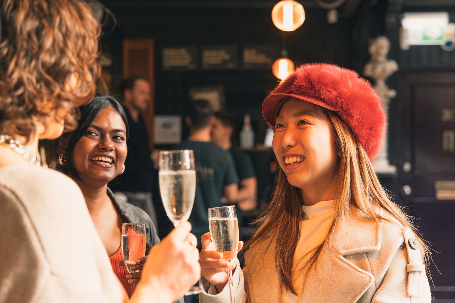 Three women smiling and clinking champagne glasses together at a bar.