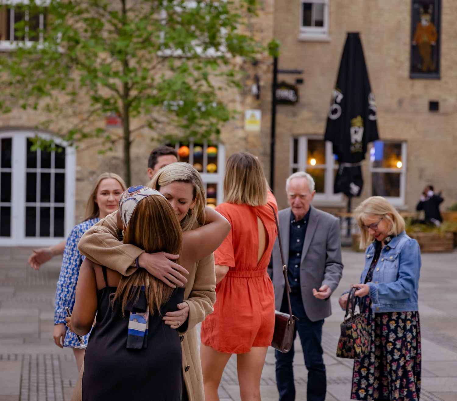 Group of people greeting and hugging each other outdoors in a brick-paved urban courtyard.