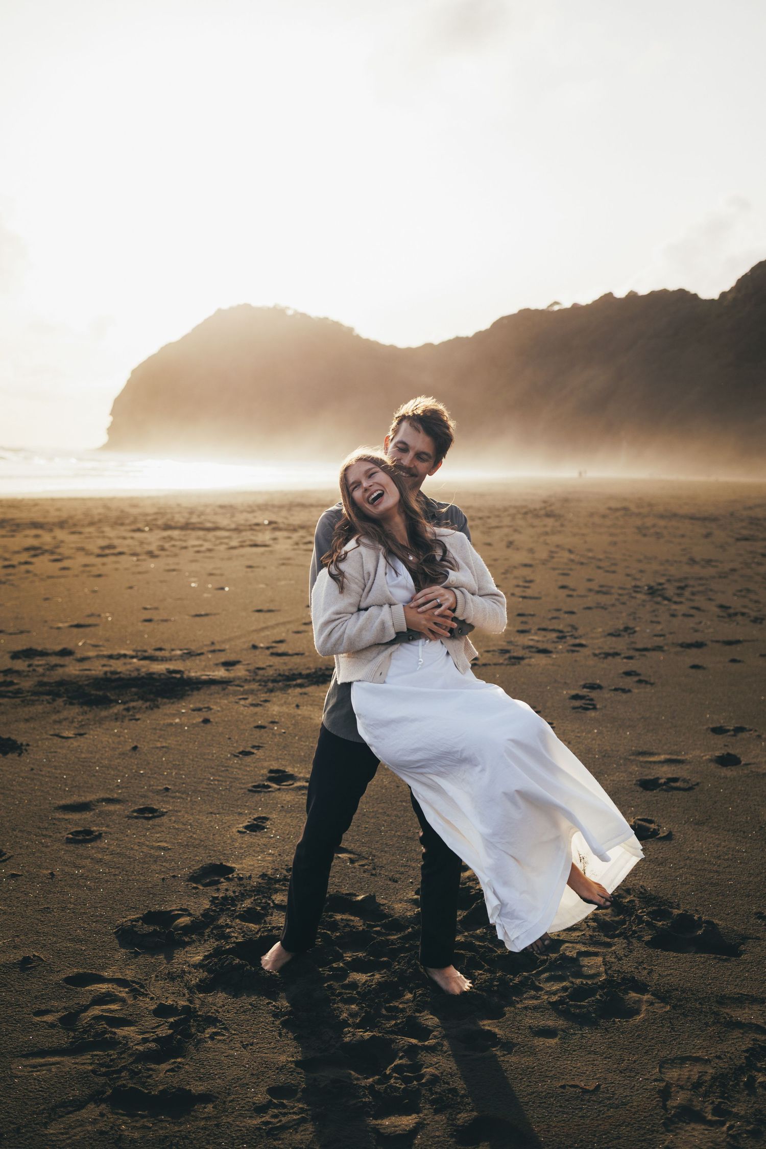 Joyful couple laughing and embracing on a dark sandy beach at golden hour with misty cliffs in the background.