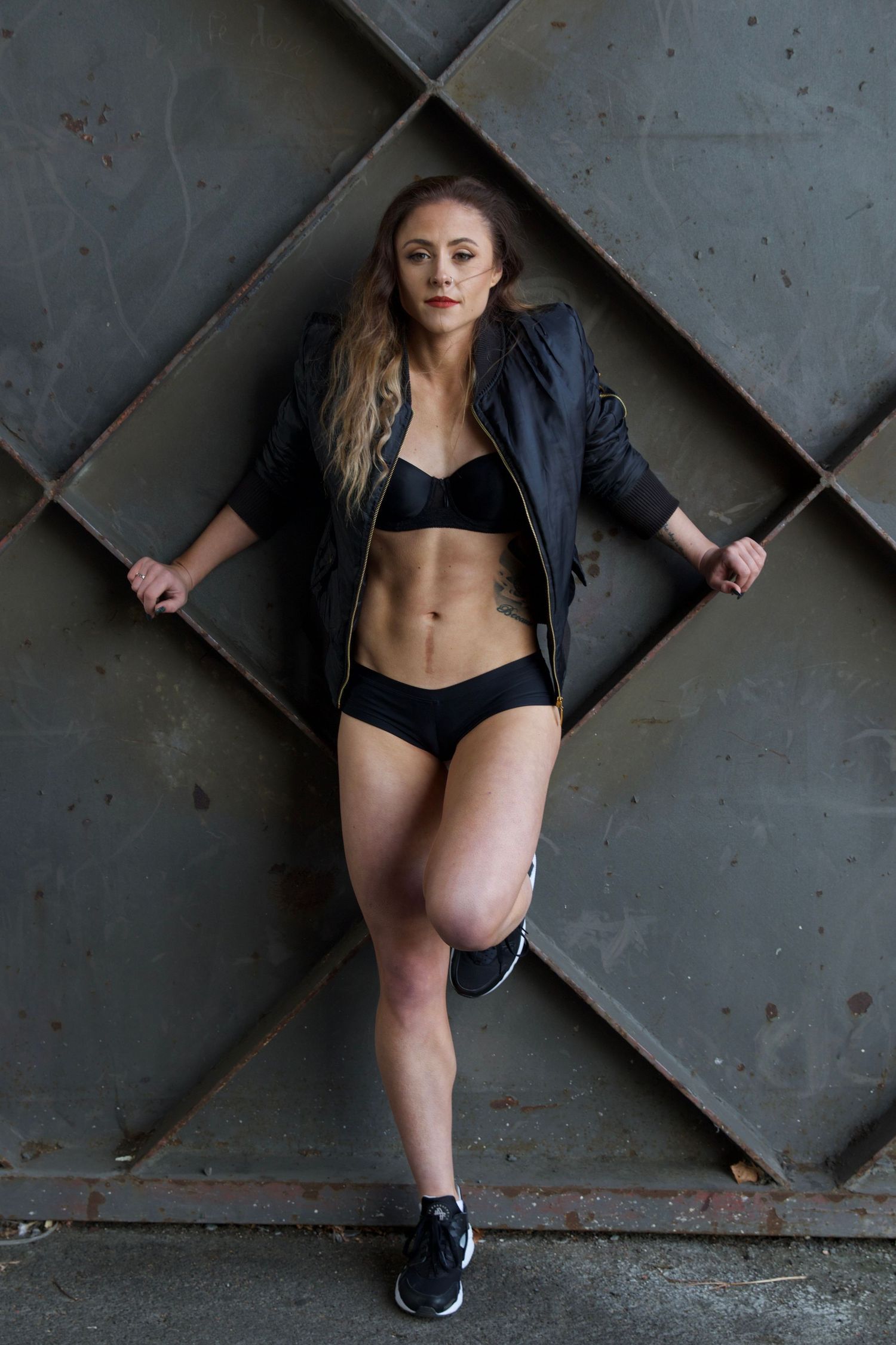 Fit blonde woman in black sports bra and shorts leaning against a metal grid wall.