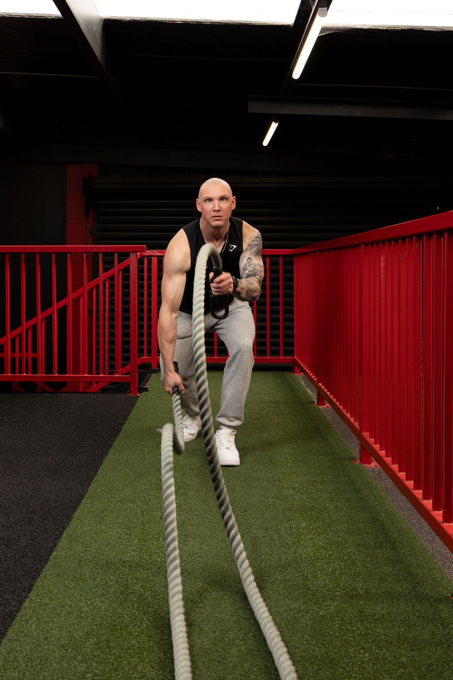 Bald tattooed man doing battle rope exercise in modern gym with red railings and green turf flooring.