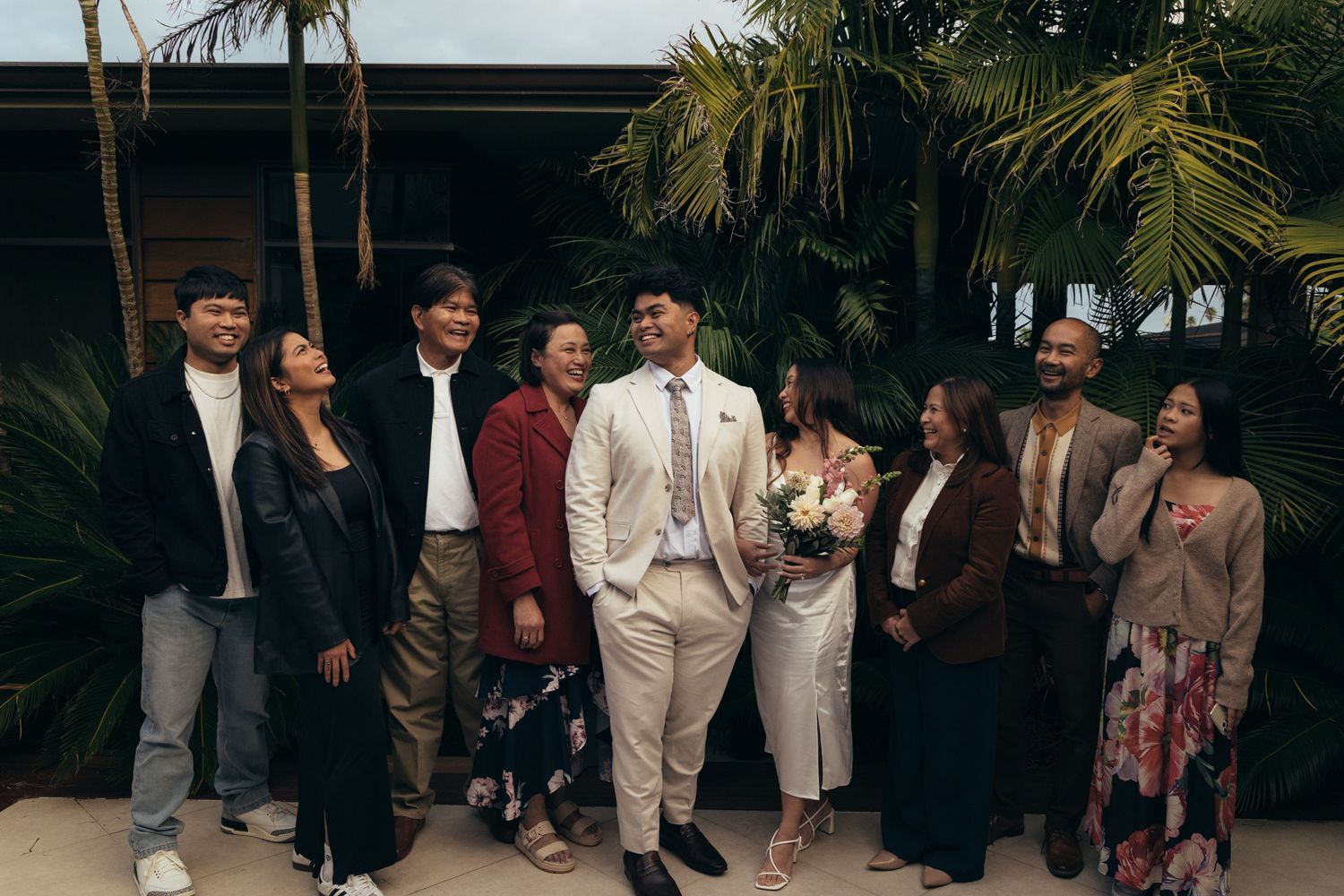 A joyful wedding party poses outdoors near palm trees, with the groom in a beige suit holding a bouquet.