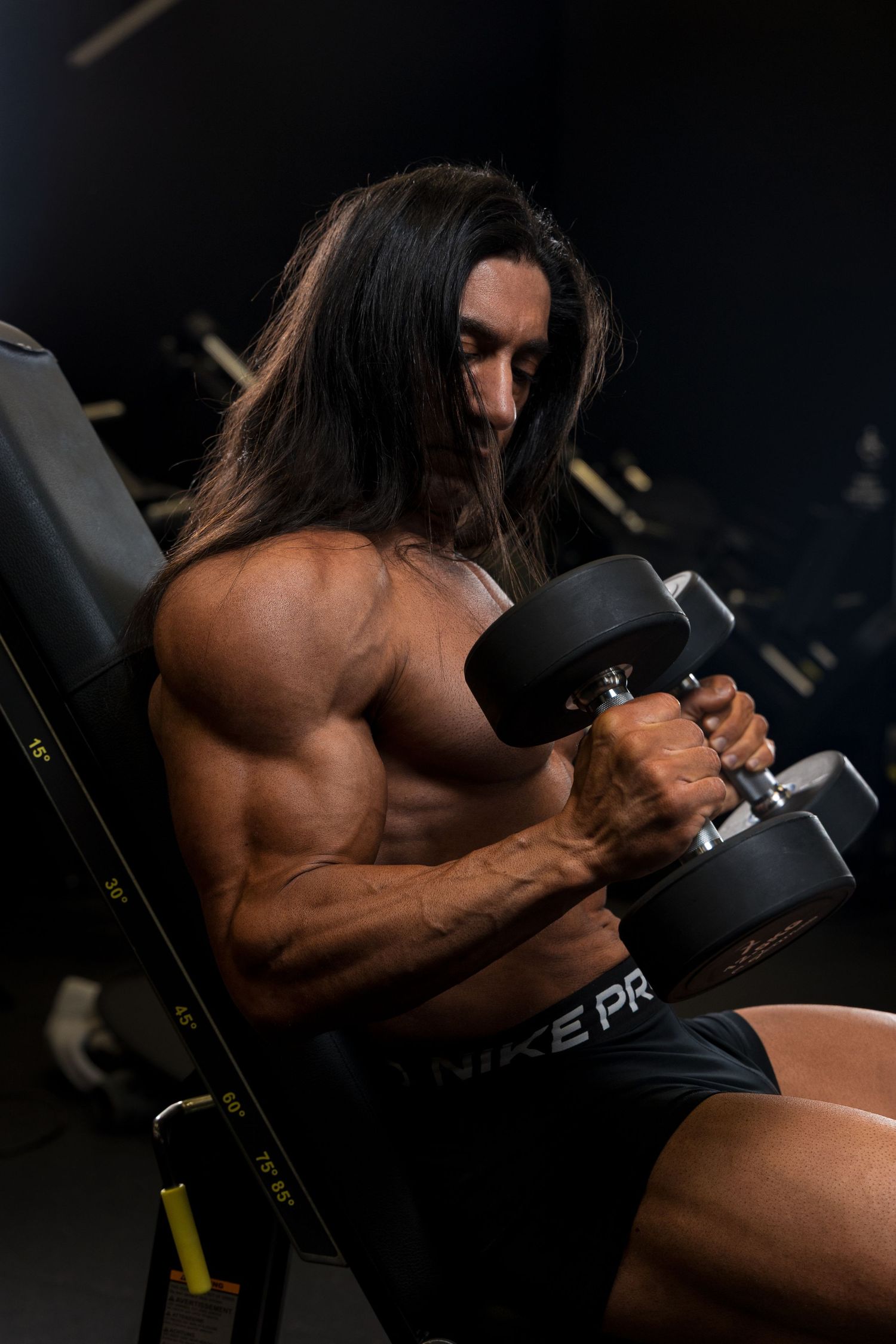 Muscular man with long hair doing dumbbell curls on a gym bench, wearing Nike Pro shorts in a dark gym.