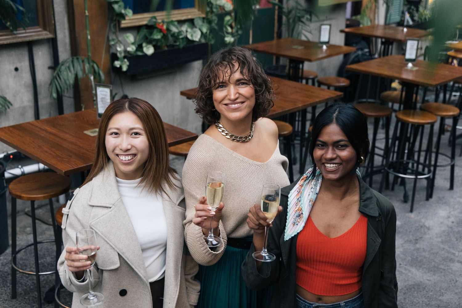 Three women smiling and holding champagne flutes while posing in a stylish indoor bar.