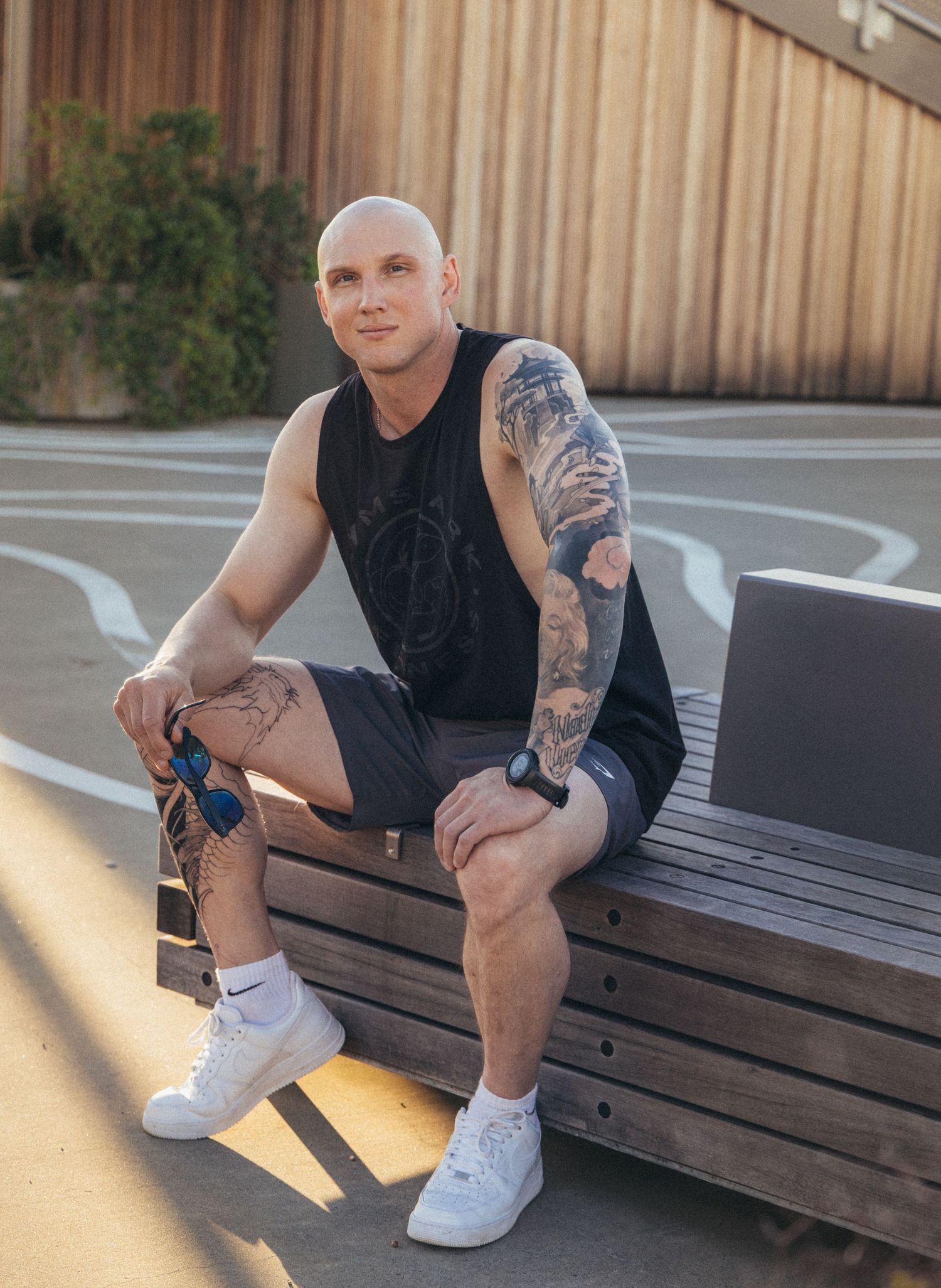 Tattooed bald man in black tank top and shorts sitting on bench at outdoor basketball court wearing white Nike shoes.